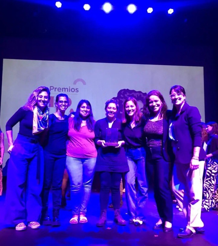 Sete mulheres estão em pé e sorrindo, enquanto a mulher central segura um prêmio.